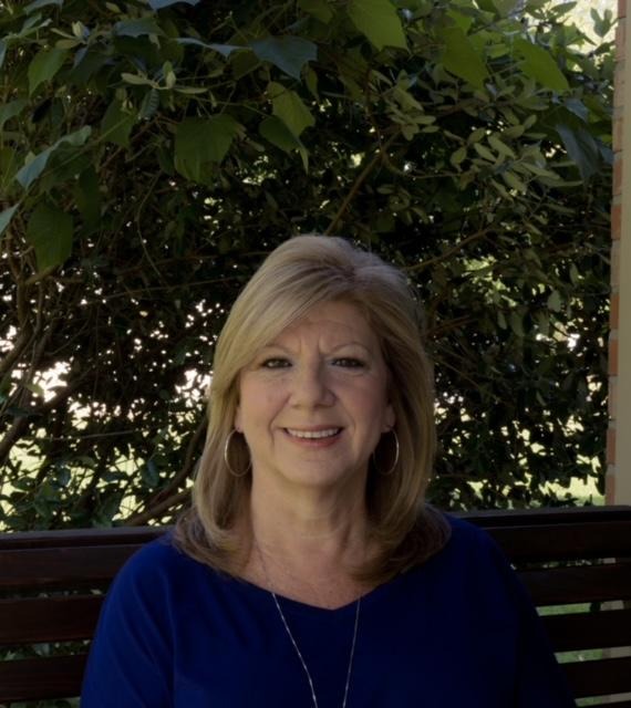 A picture of Mrs. Patty Rogge sitting on a bench in front of a large bush.